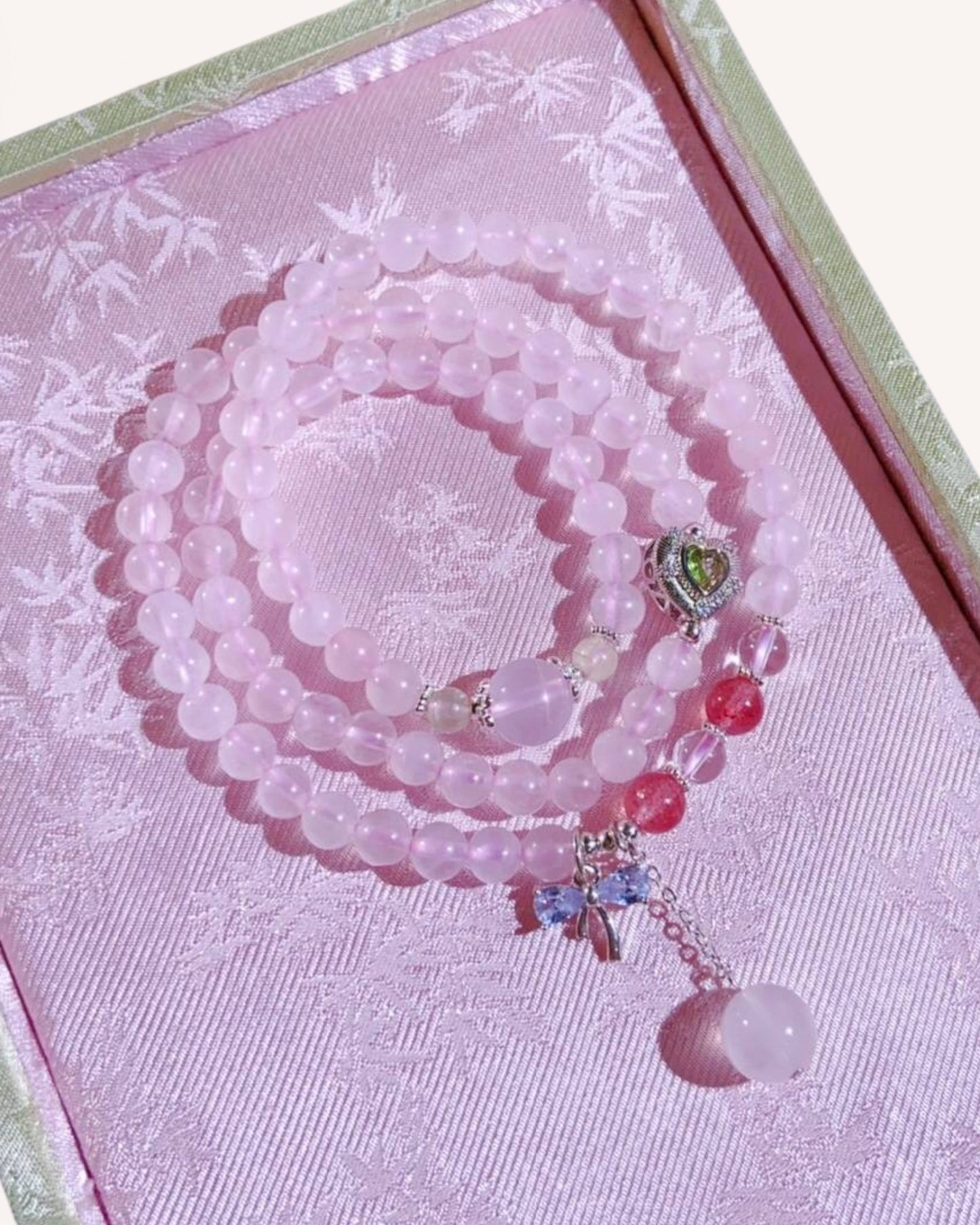 Triple-layer rose quartz bracelet with strawberry quartz, grape stone, and heart charm, styled on a pink silk jewelry box for love and positive energy.