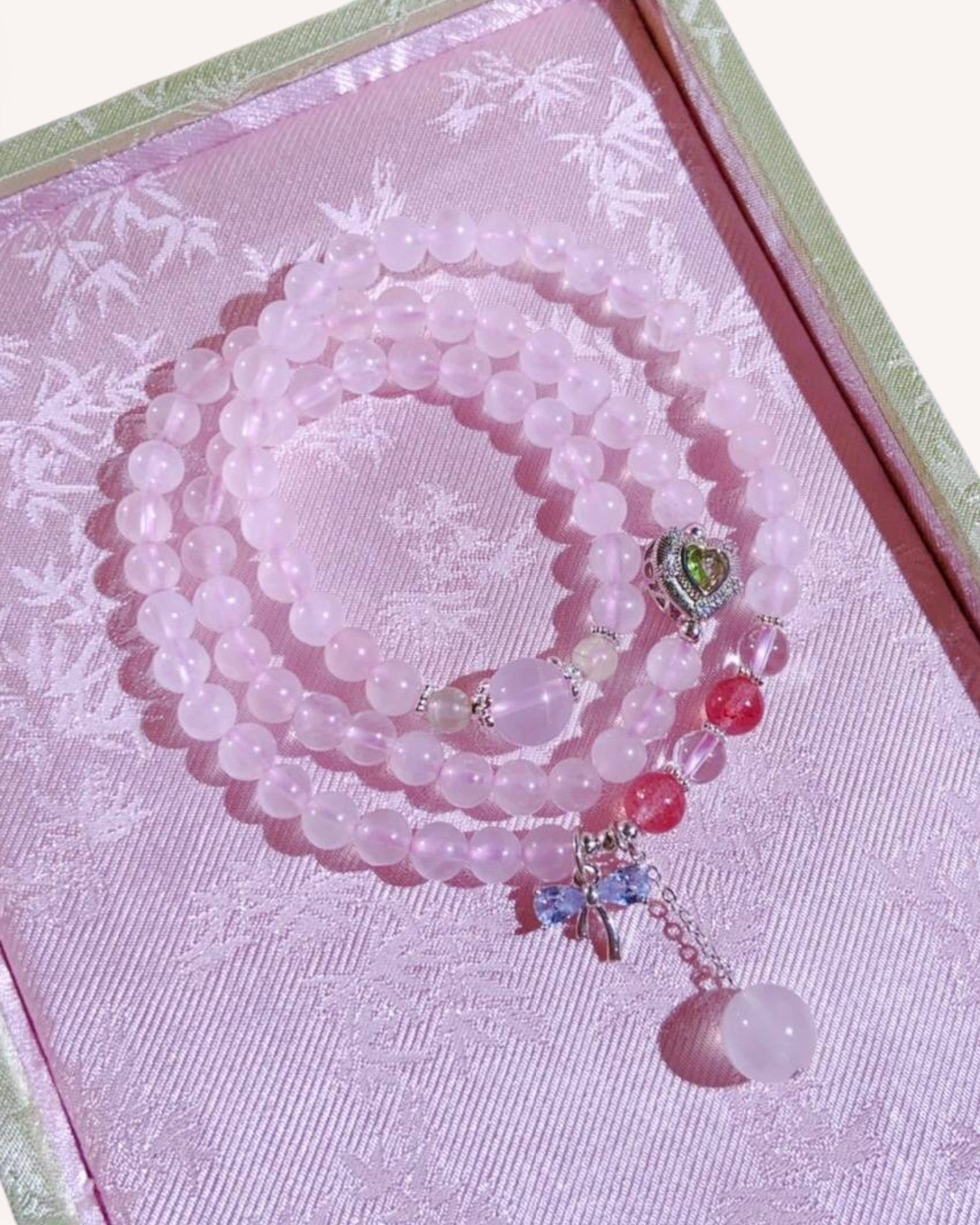 Triple-layer rose quartz bracelet with strawberry quartz, grape stone, and heart charm, styled on a pink silk jewelry box for love and positive energy.