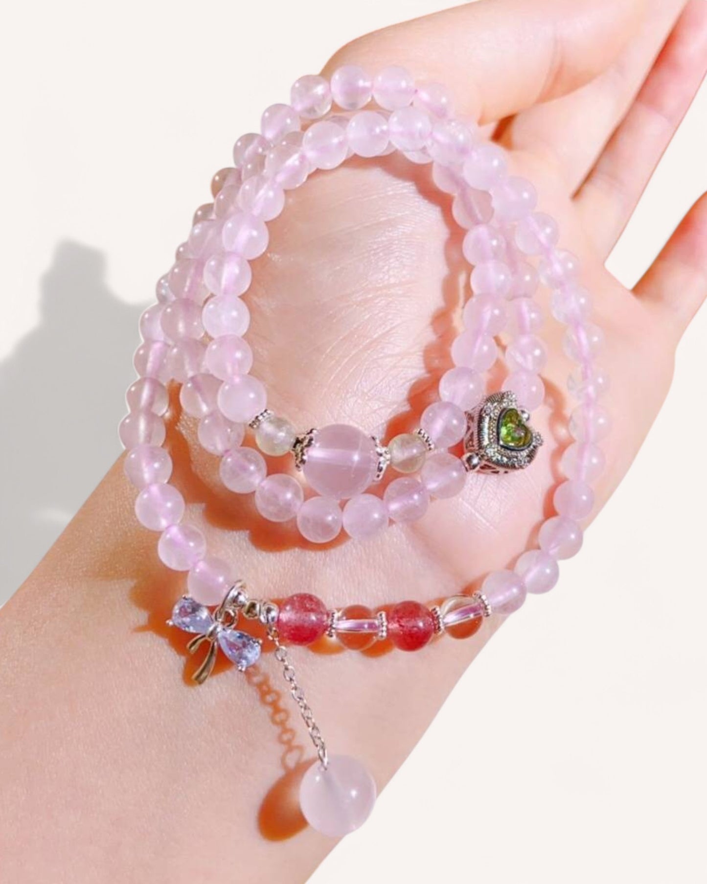Hand wearing triple-layer rose quartz and strawberry quartz bracelet with dangling heart charm, designed for love, happiness, and emotional well-being.